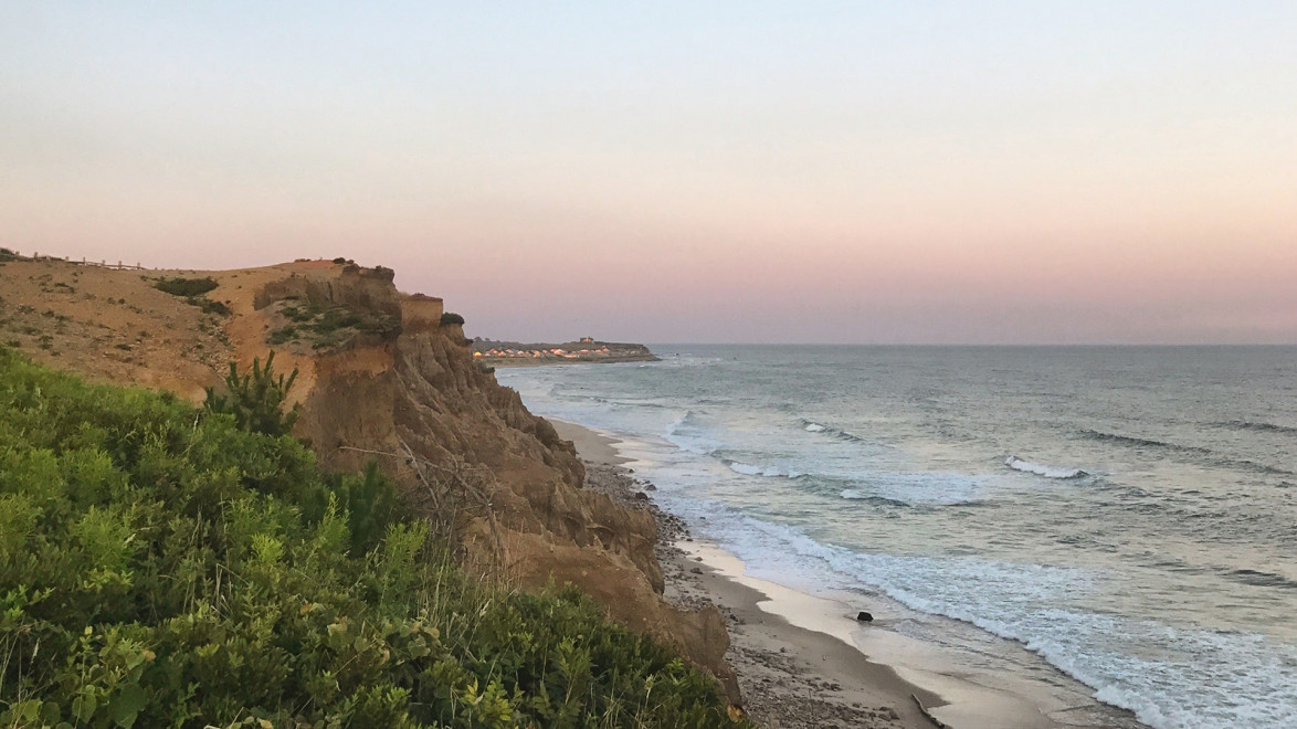 Coastal cliffside at sunset with green vegetation, sandy beach, and waves gently lapping against the shore.
