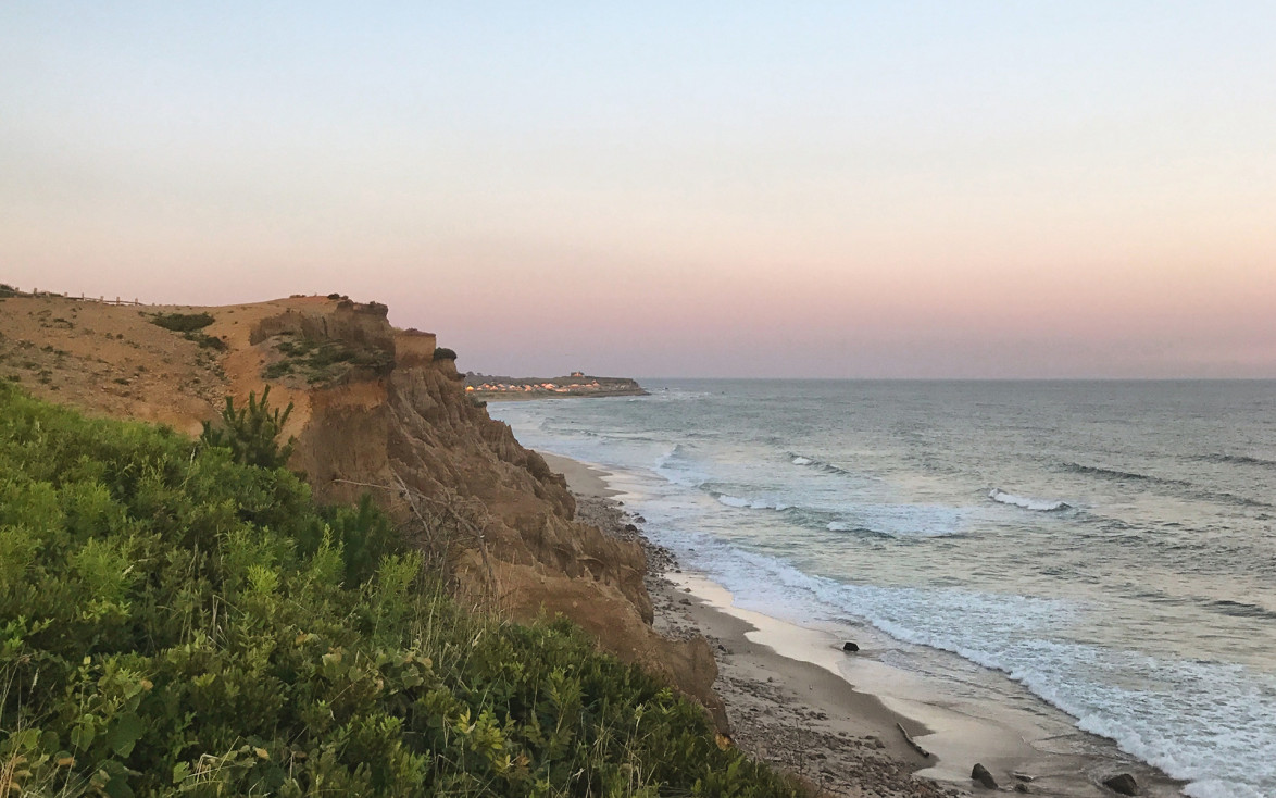 Coastal cliffside at sunset with green vegetation, sandy beach, and waves gently lapping against the shore.