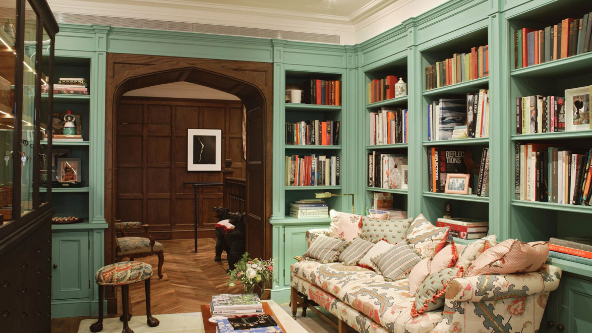 Cozy library with green bookshelves, a patterned sofa, wooden furniture, and an arched wooden doorway in the background