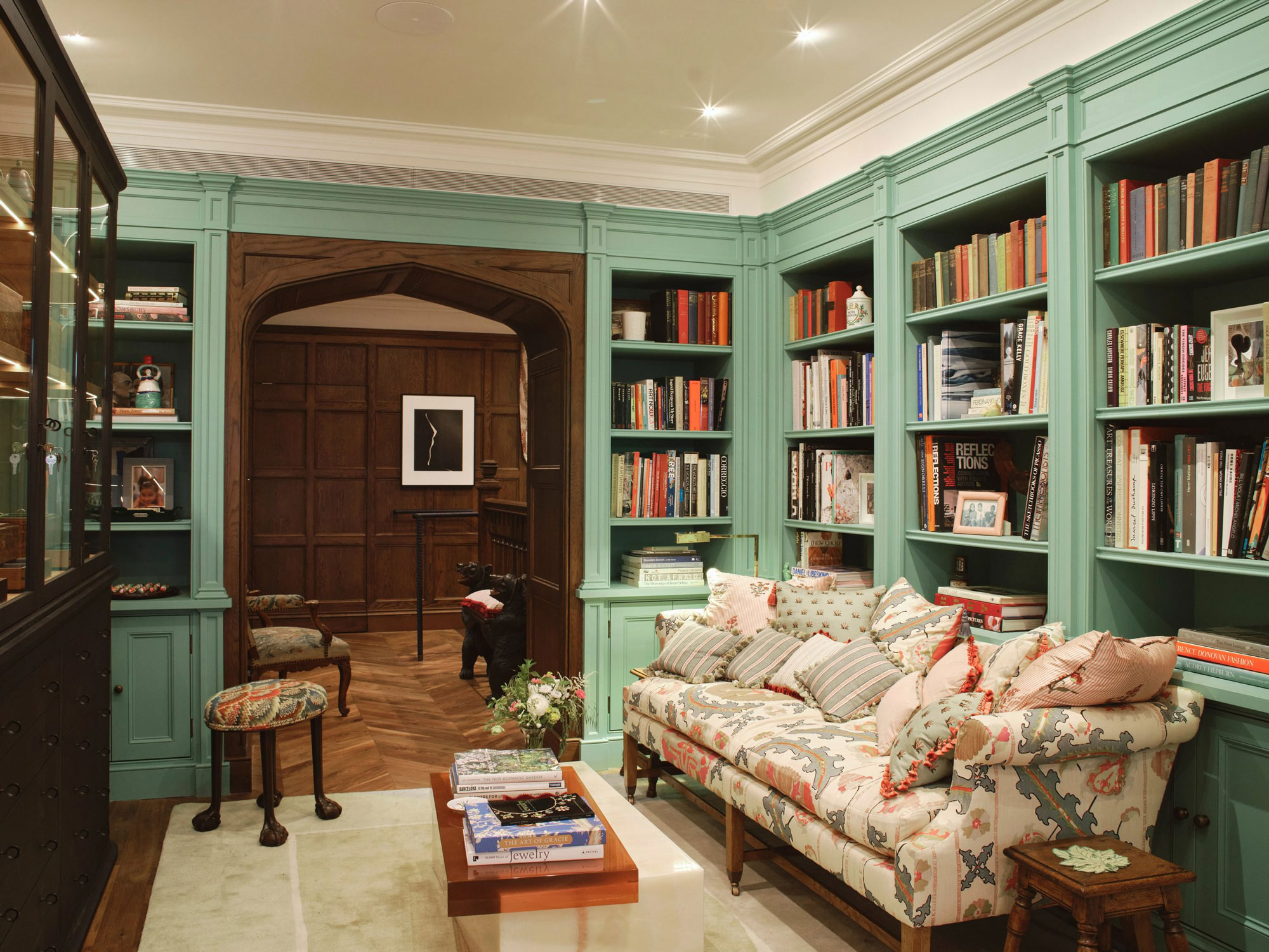 Cozy library with green bookshelves, a patterned sofa, wooden furniture, and an arched wooden doorway in the background