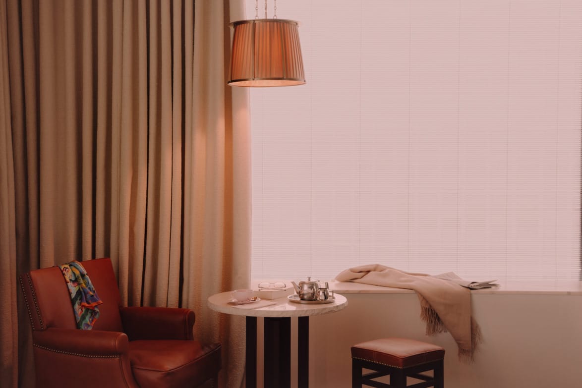 Cozy room with a leather chair, small round table set for tea, and a window with blinds filtering warm light.