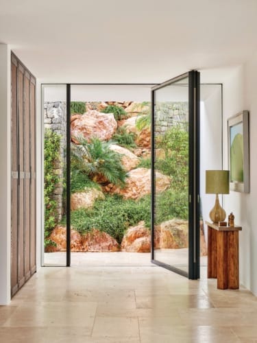 Open glass doors leading to a rocky garden with lush greenery, seen from a modern room with a wooden console and lamp.