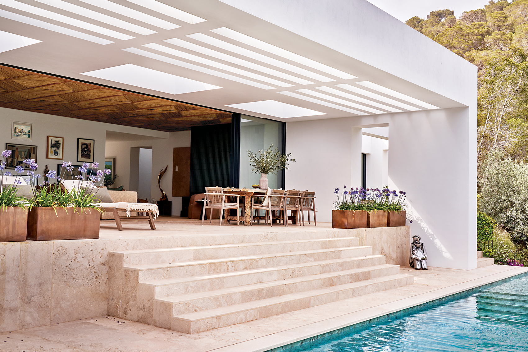 Modern open-air living room with dining set, surrounded by greenery, leads to a pool with decorative planters on steps.