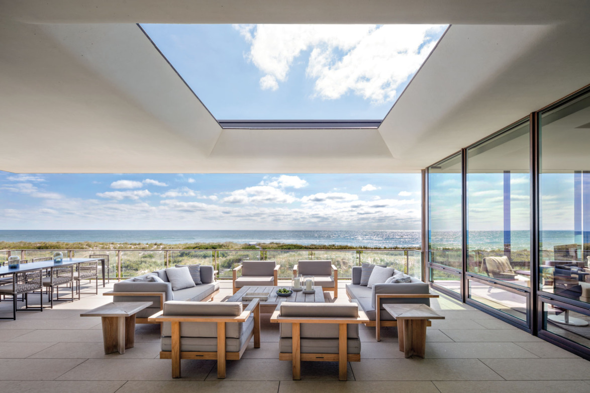 Modern outdoor patio with ocean view, featuring contemporary furniture and a large glass window.