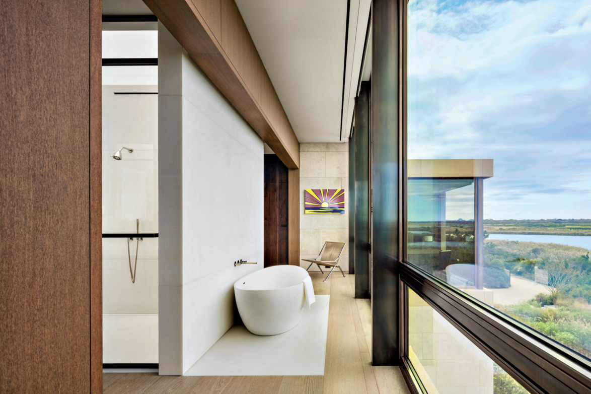 Modern bathroom with a freestanding white tub, large windows offering scenic views, and a colorful painting on the wall.