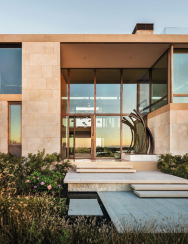 Modern house facade with large glass windows and stone walls, surrounded by landscaping and a clear blue sky in the background.
