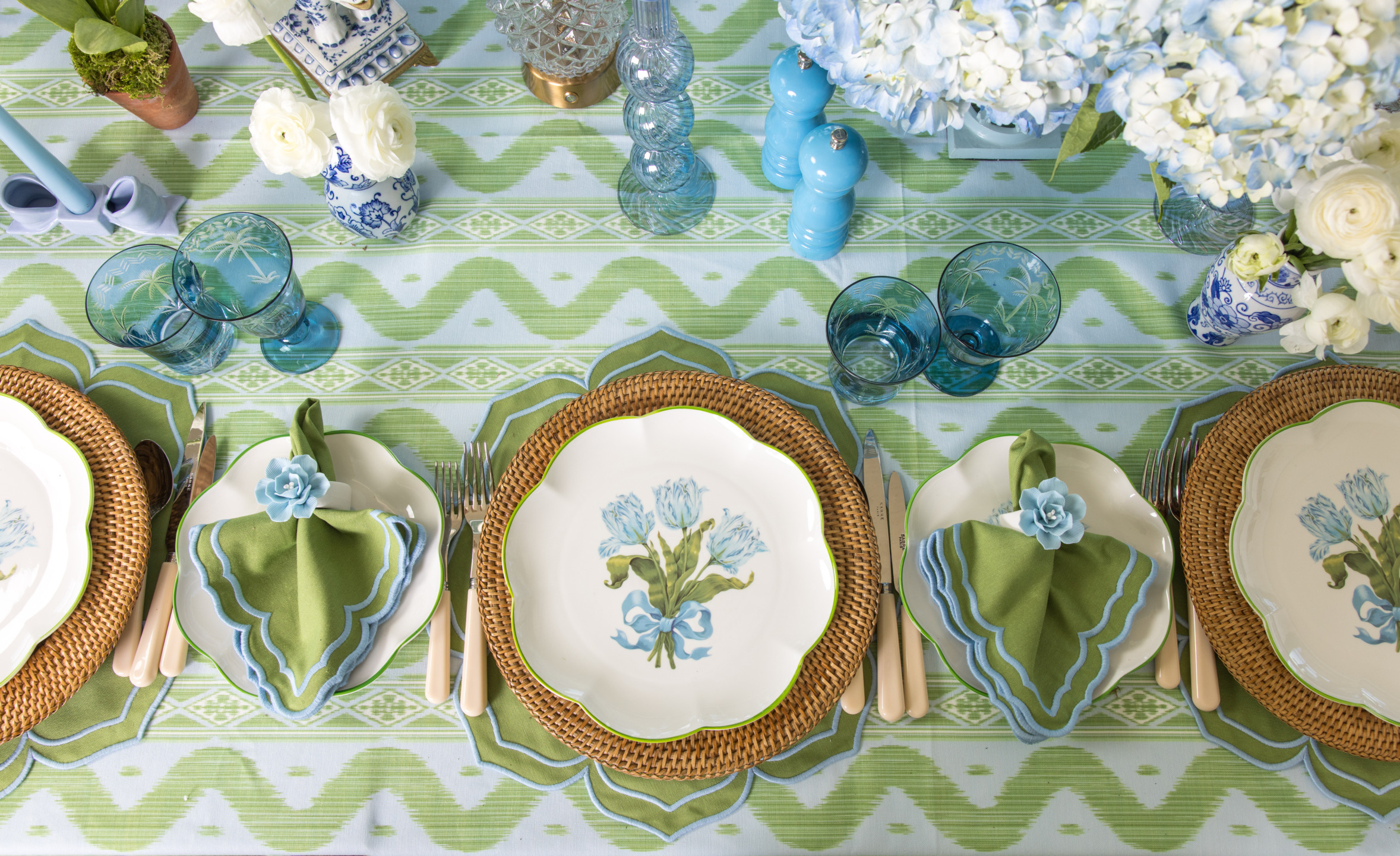 Elegant table setting with floral plates, green napkins, and blue glassware on a green and white patterned tablecloth.