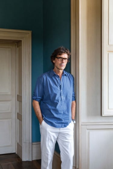 Man in blue shirt and white pants standing by a wall with hands in pockets in a room with teal and wooden accents