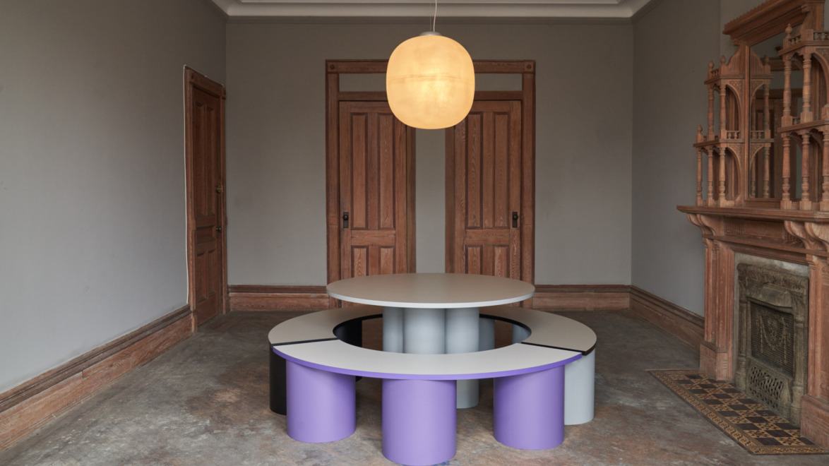 Modern room with circular table and benches, purple accents, wooden doors, ornate fireplace, and a glowing ceiling light.