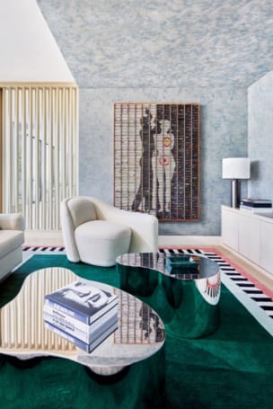 Modern living room with abstract art, white chairs, and reflective green coffee tables on a green rug with books stacked.