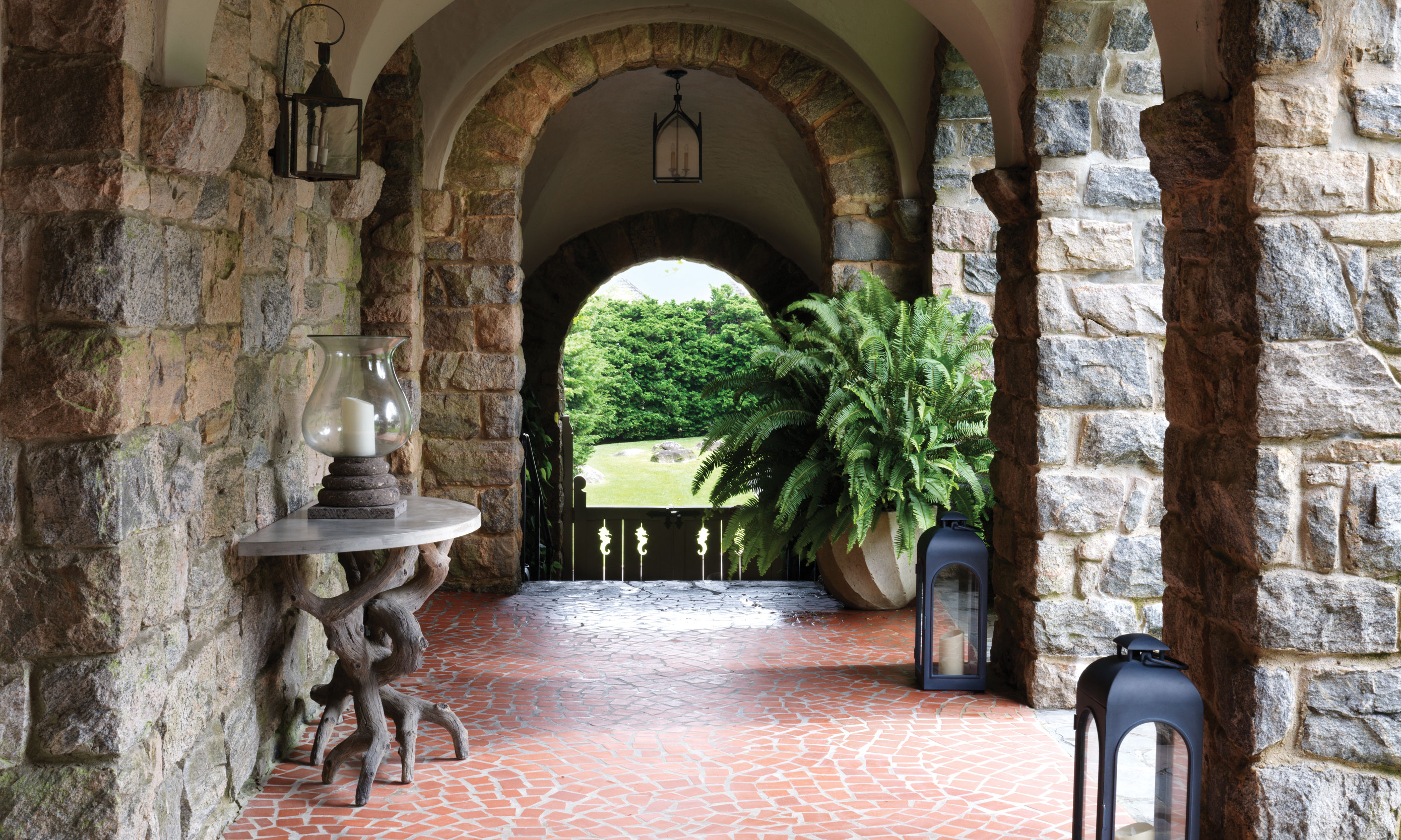 Stone archway with brick floor, lanterns, table with vase, and lush greenery in the background.