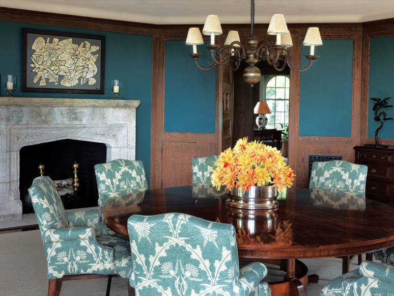 Elegant dining room with floral-patterned chairs, round wooden table, chandelier, fireplace, and a vase of yellow flowers.
