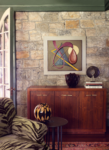Abstract art on stone wall above wooden cabinet with decorative items, patterned armchair, and round side table.