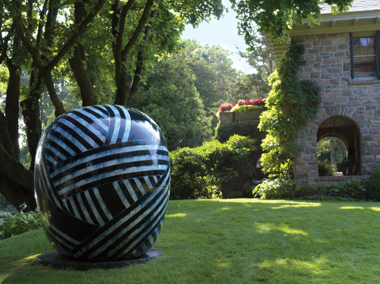 Large striped spherical sculpture in a lush garden setting with trees and a stone building in the background.