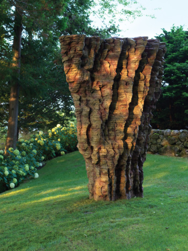 Tall stone sculpture with a textured surface, set on a lush green lawn surrounded by trees and flowering bushes.