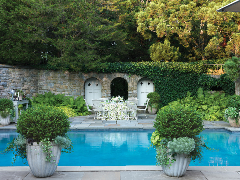 Poolside dining area with a table set for a meal, surrounded by greenery and a stone wall with arches in the background.