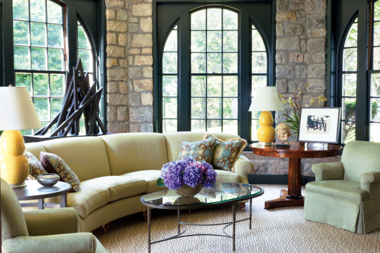 Cozy living room with stone walls, large windows, curved sofa, glass coffee table, and yellow lamps on a side table.