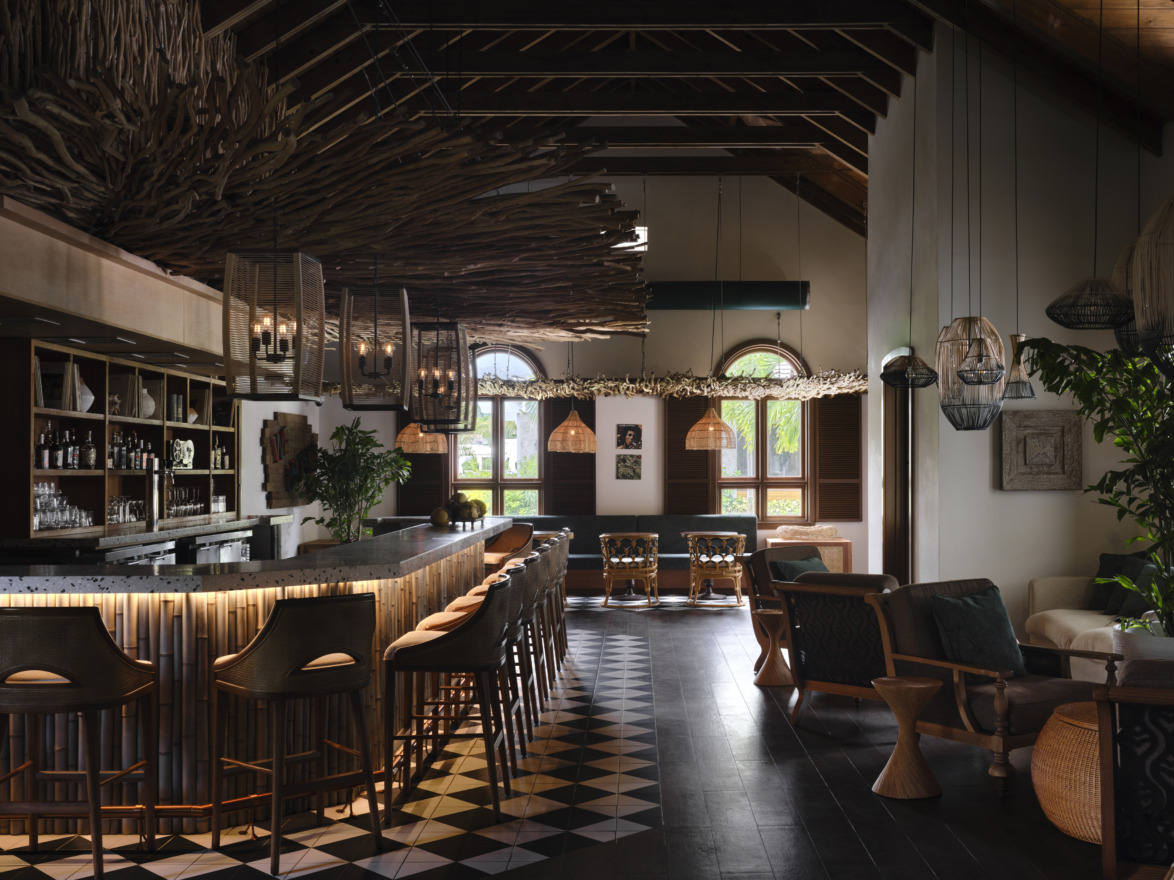 Rustic restaurant interior with a wooden bar, stools, woven light fixtures, and large windows with natural light.