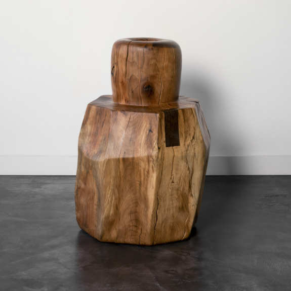 Carved wooden stool with a natural finish, featuring a cylindrical top and octagonal base, placed on dark concrete floor.