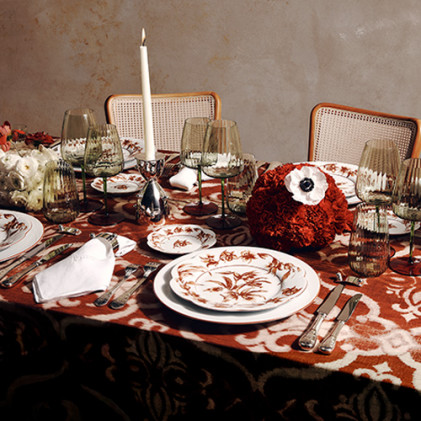 Elegant table setting with floral-patterned plates, green glasses, red floral centerpiece, and a tall candle on a patterned cloth.