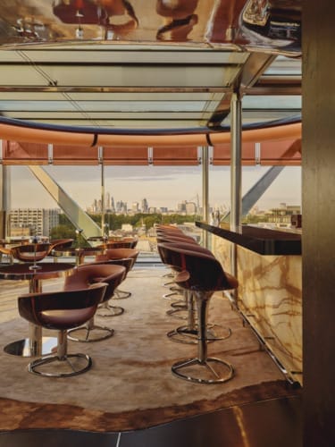 Modern bar interior with high stools, marble counter, and city skyline view through large windows, during sunset.