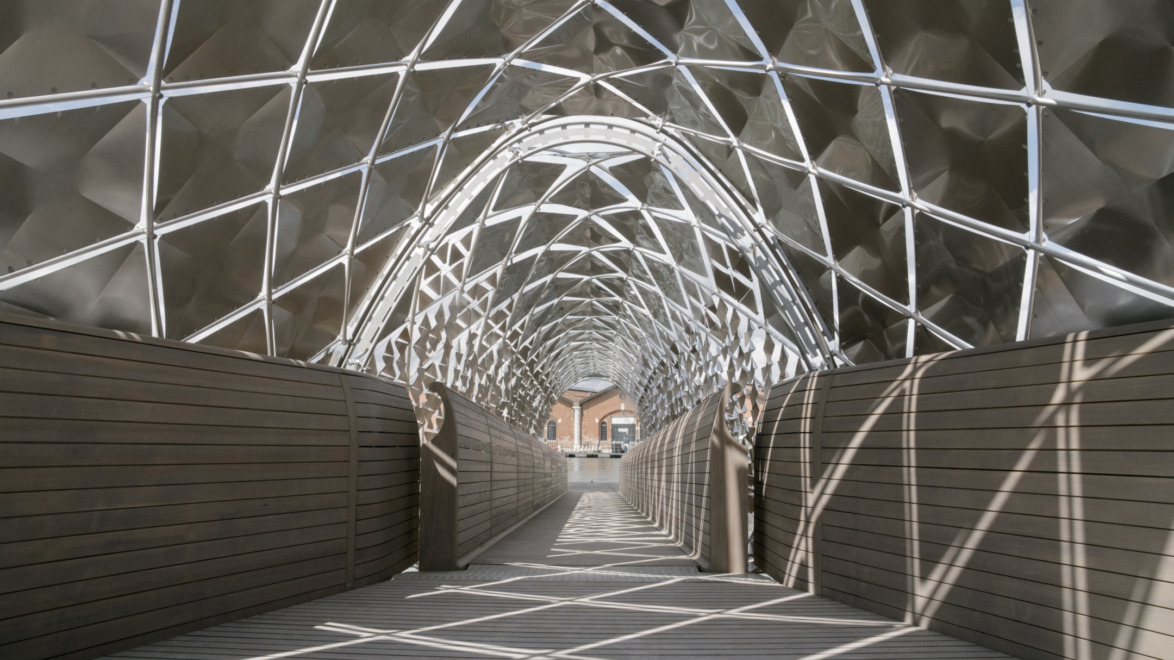 Modern geometric bridge with metal lattice roof and wooden walkway, casting intricate shadows.