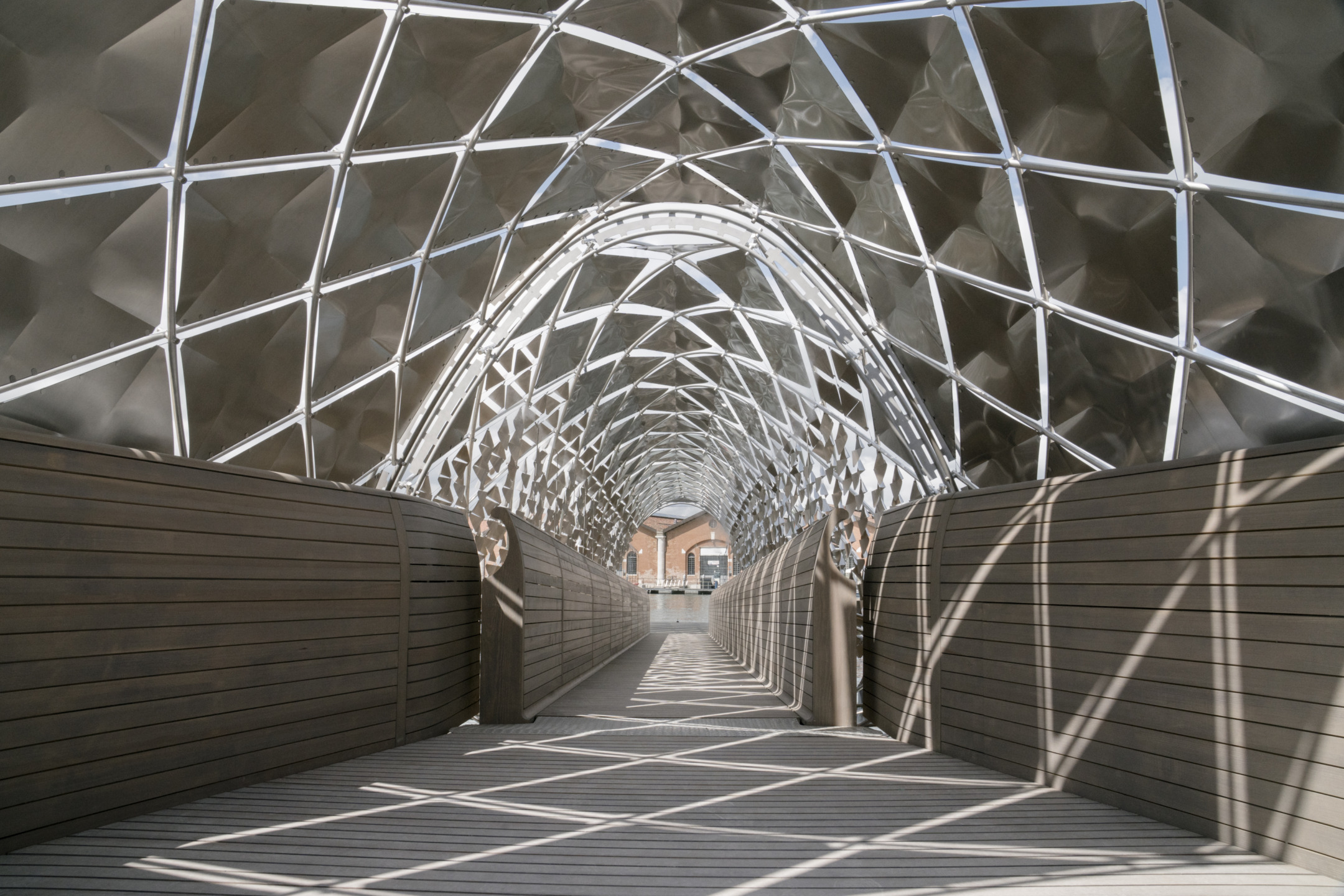 Modern geometric bridge with metal lattice roof and wooden walkway, casting intricate shadows.