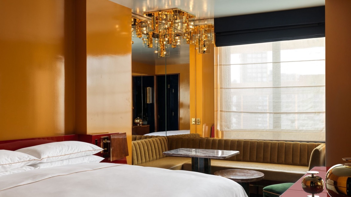 Modern hotel room with yellow walls, a luxurious bed, stylish chandelier, and a window with sheer curtains and a city view.