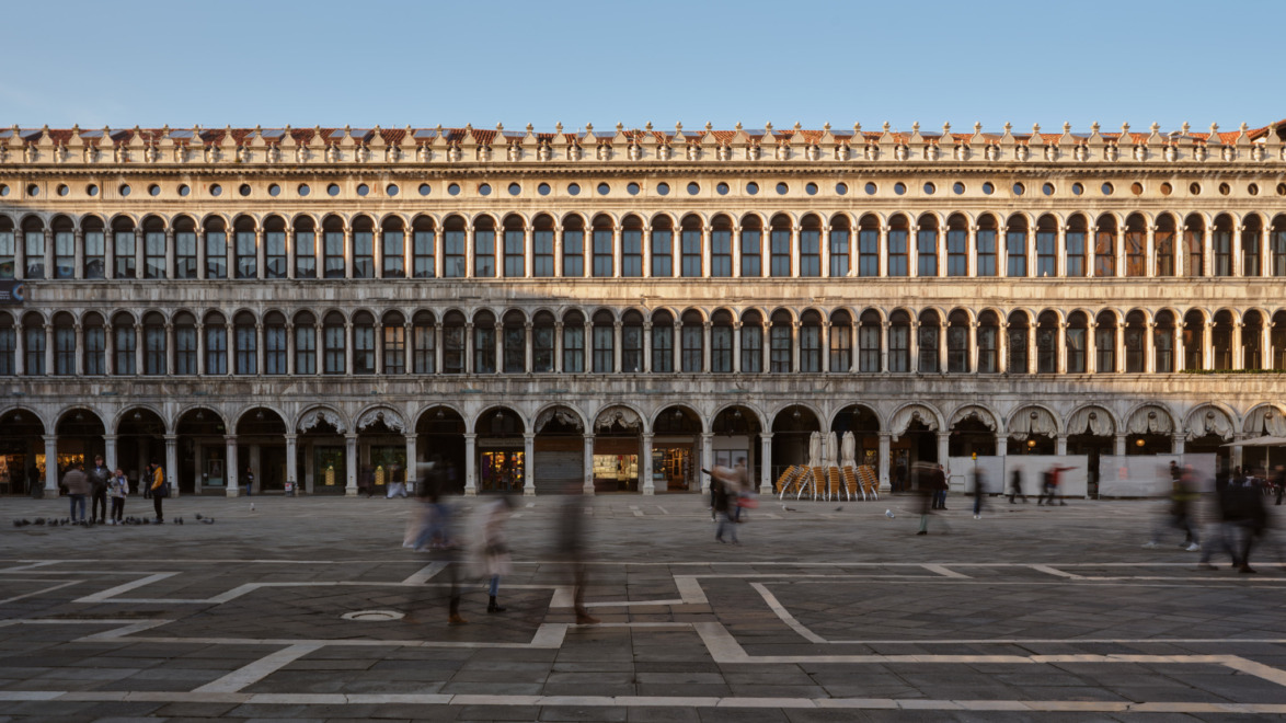 Historic building in a spacious square with people walking, under a clear blue sky.