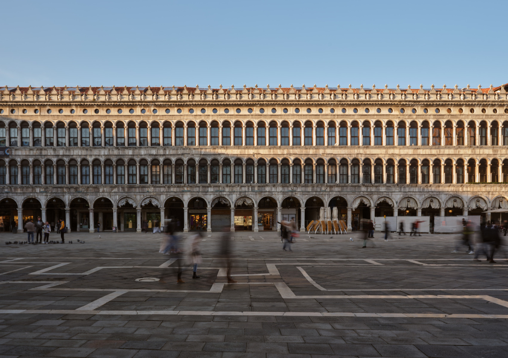 Historic building in a spacious square with people walking, under a clear blue sky.