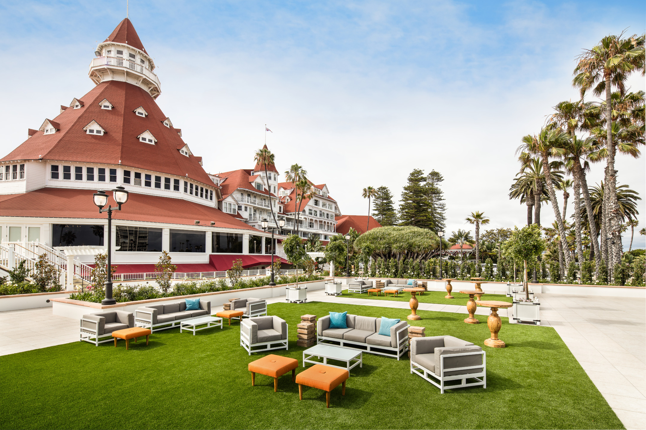 Outdoor seating area with modern furniture on grass, surrounded by palm trees, near a large red-roofed historic building.