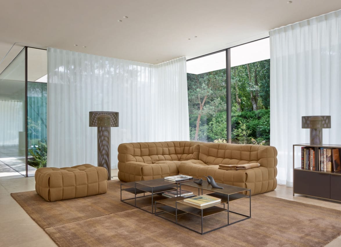 Modern living room with large windows, beige sectional sofa, matching ottoman, glass coffee table, and sheer white curtains.