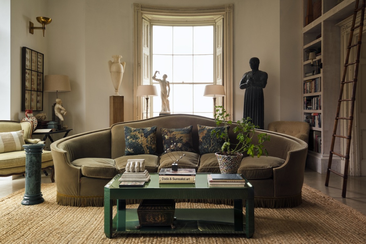Cozy living room with green sofa, decorative statues, bookshelves, and a coffee table with plants and books.