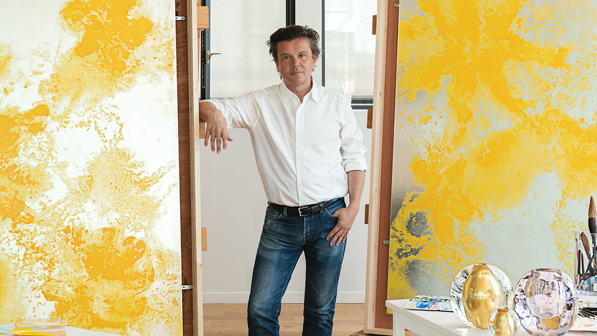 Artist standing in a studio surrounded by abstract yellow paintings, wearing a white shirt and jeans, with art supplies nearby.