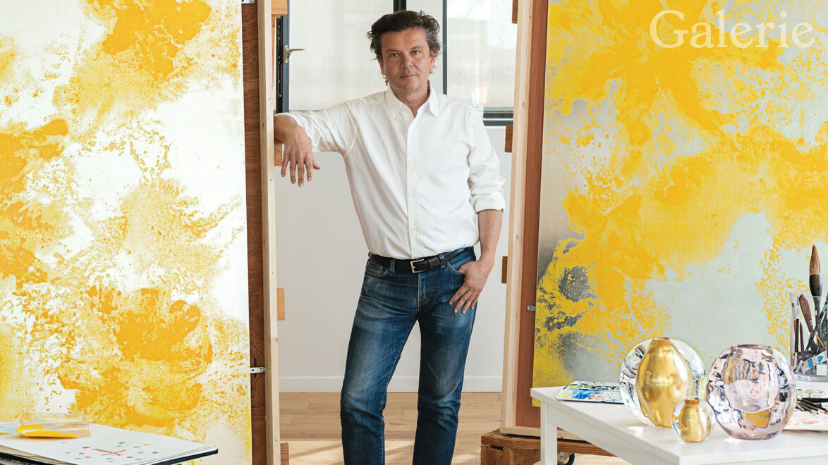 Person standing in art studio with abstract yellow paintings, wearing a white shirt and jeans, surrounded by art supplies.