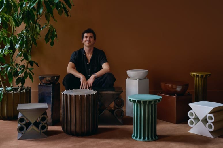 Man sitting among various modern, artistic stools and tables in a stylish room with warm lighting and a large plant.
