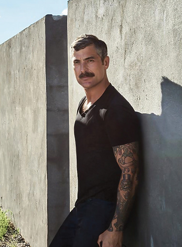 Man with tattoos and a mustache standing against a concrete wall in a black shirt, looking into the camera.