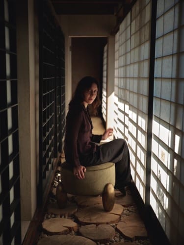 Woman sitting on a stool in a narrow corridor with shoji screens, bathed in soft light, wearing a dark outfit, looking serene.