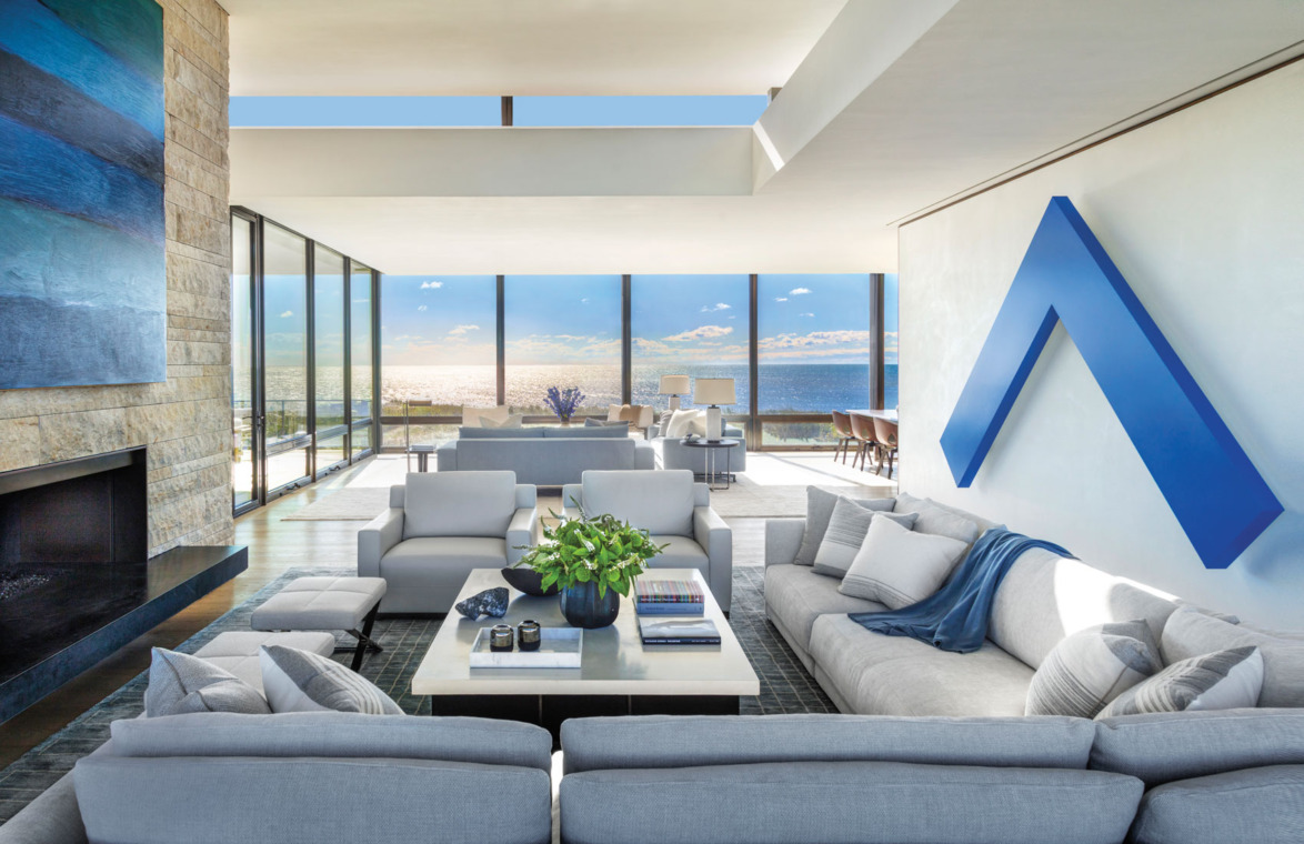 Modern living room with large windows, ocean view, gray sofas, blue wall art, and a central coffee table with plants.