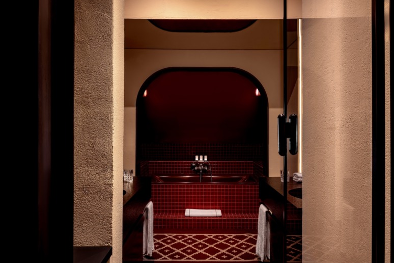 Elegant modern bathroom with red tiles, arched mirror, and soft lighting, featuring a dark wooden counter and towels.