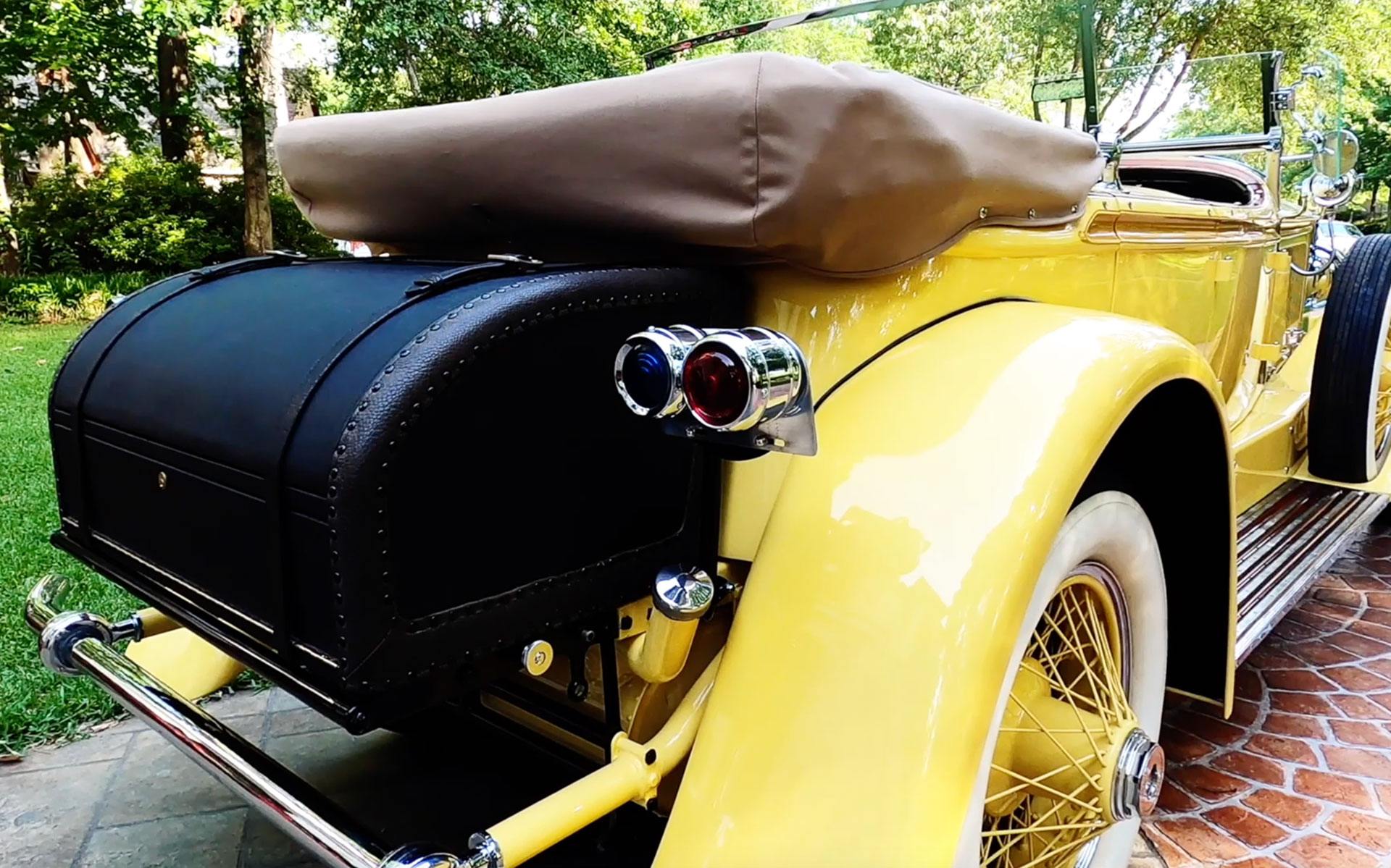 Yellow vintage car with black trunk, beige roof, and classic details parked on a brick driveway surrounded by trees.