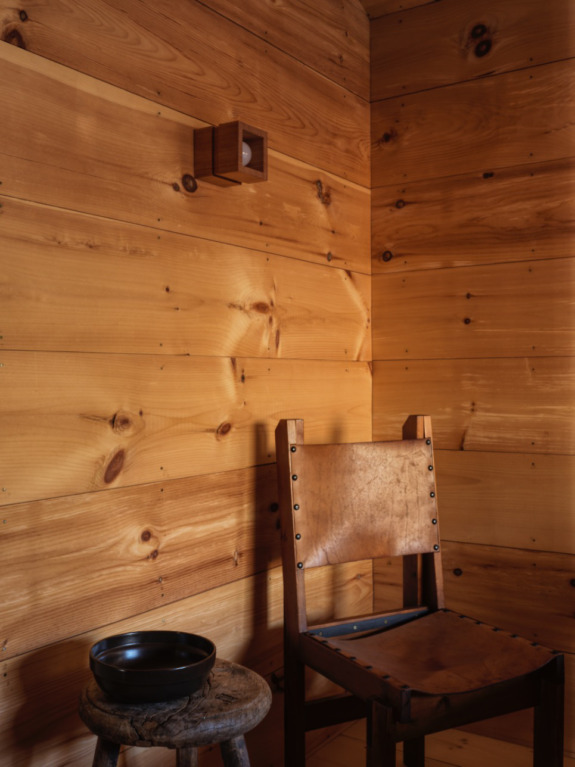 Wood-paneled room corner with a leather chair, small stool, black bowl, and a cube-shaped wall sconce.