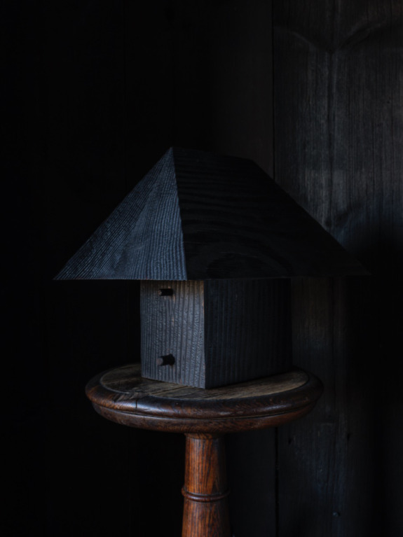 Dark wooden birdhouse with a triangular roof, positioned on a wooden stool against a dark background.