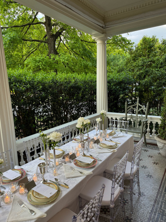 Elegant outdoor table setting with white linens, candles, and flowers on a porch surrounded by greenery.