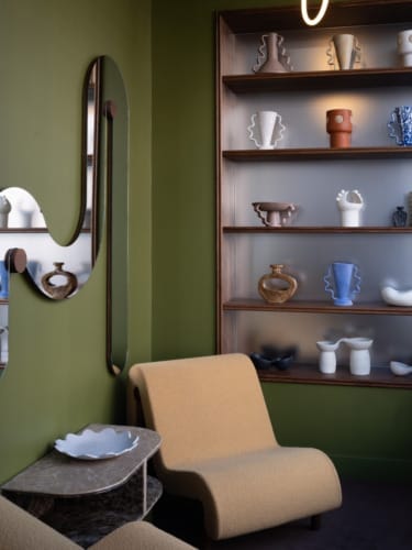 Modern room with green walls, sculpted vases on shelves, large mirror, beige chairs, and a small marble side table.