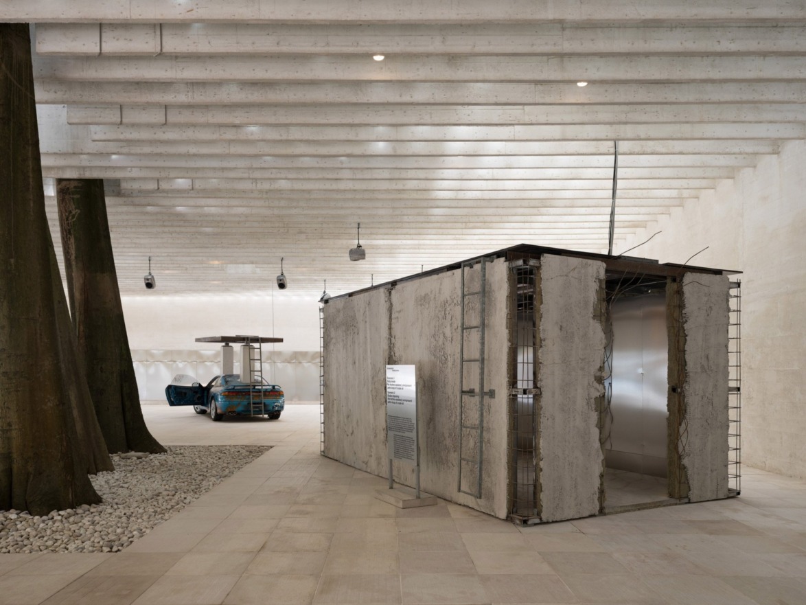 Modern art installation in a spacious gallery with trees, a weathered structure, and a partially covered car in the background.