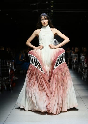 Model walking runway in a unique pleated gown with pink and white hues, featuring artistic cage-like elements at the hips.