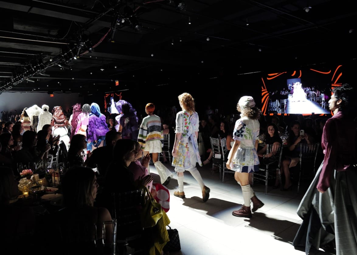 Models walking down a dimly lit fashion runway with audience seated on both sides.