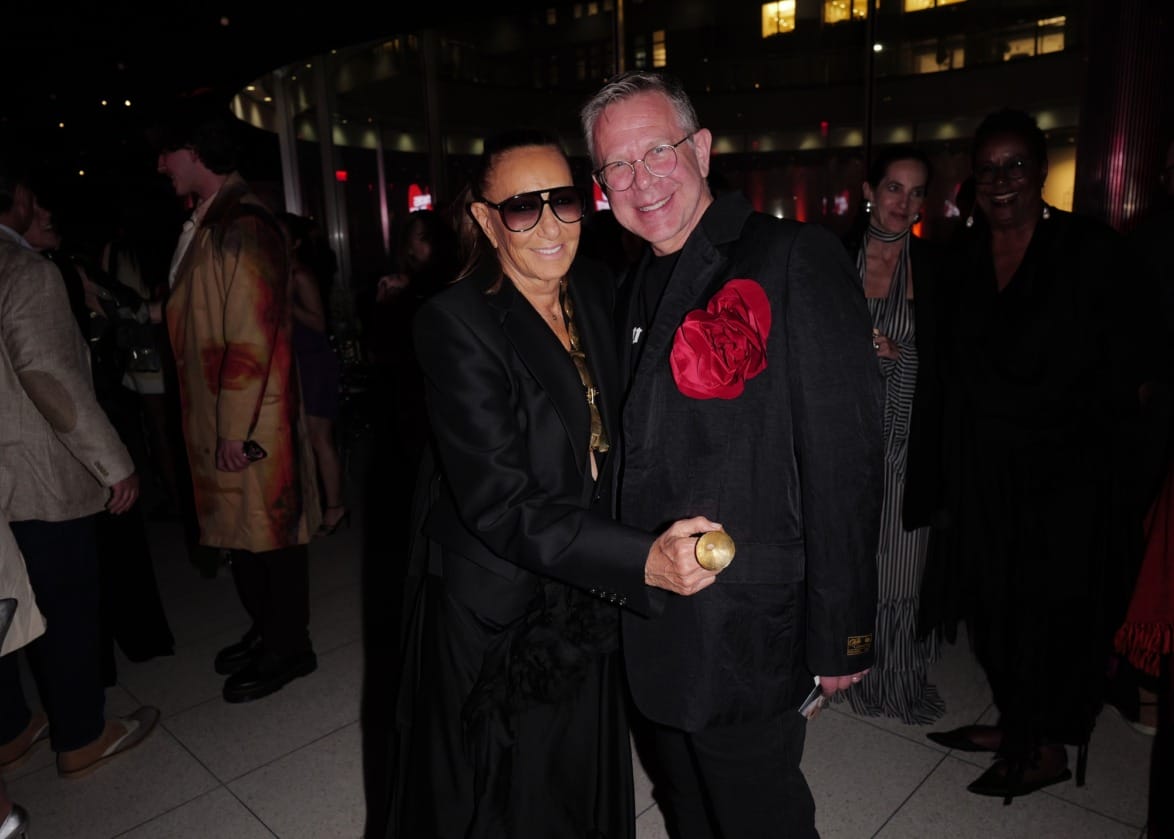 Two people posing and smiling at an indoor event, surrounded by other attendees and warm lighting.