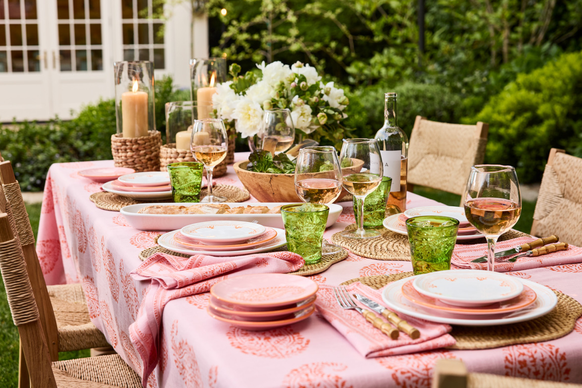Elegant outdoor table setting with pink tablecloth, wine glasses, decorative plates, and candles in a garden setting.